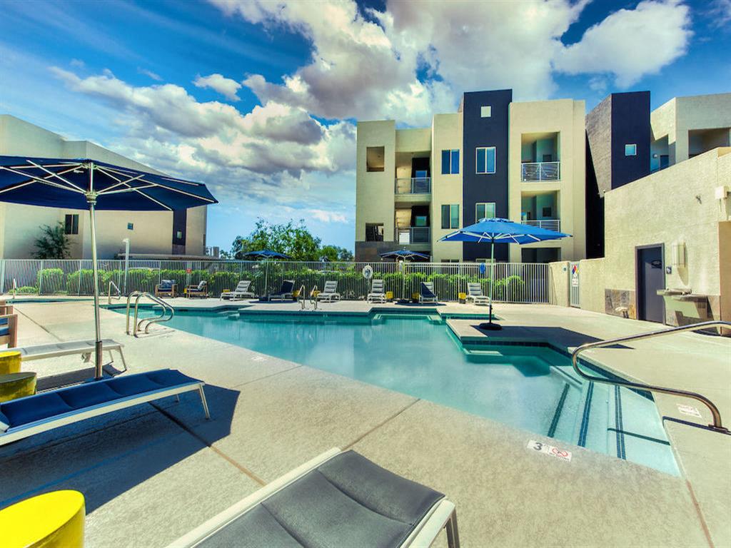 Pool with buildings in the background and patio umbrella up front. Four lounge chairs line the pool.