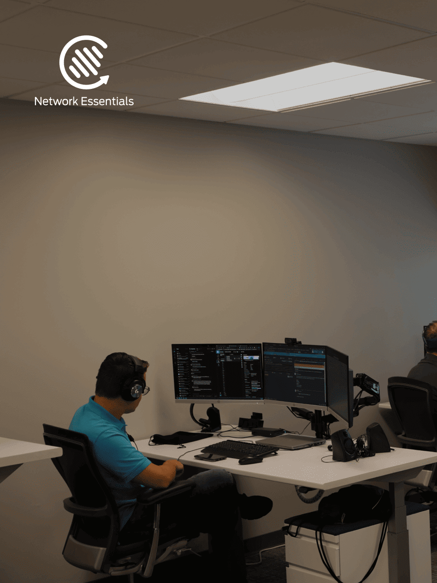 A man in a blue shirt and headphones is sitting at a desk with two computer monitors in an office environment. The logo for 'Network Essentials' is visible in the upper left corner.
