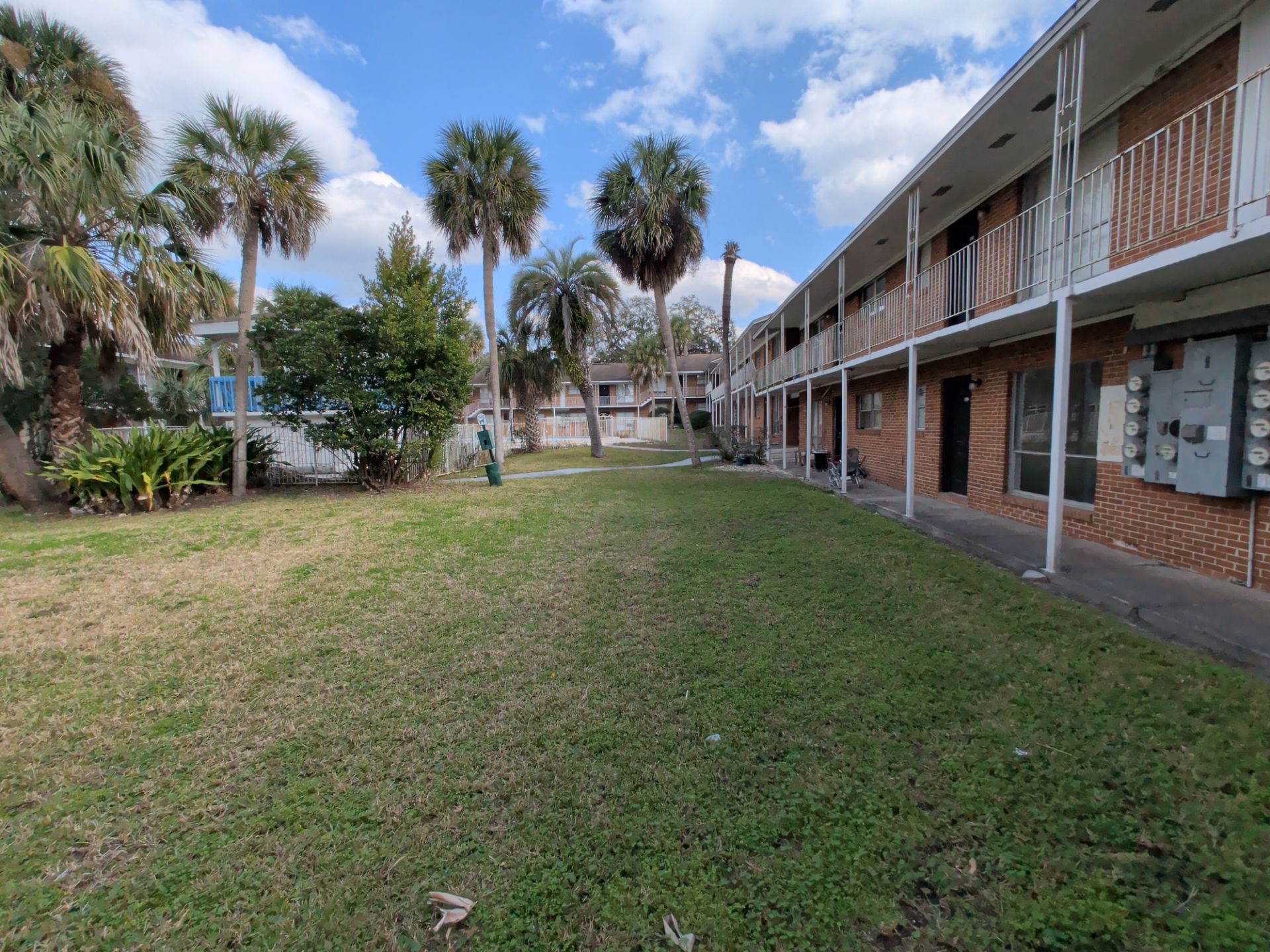 Apartment complex courtyard with grassy lawn, palm trees, and landscaped greenery. Two-story brick residential buildings with exterior walkways and balconies surround the shared outdoor community space.