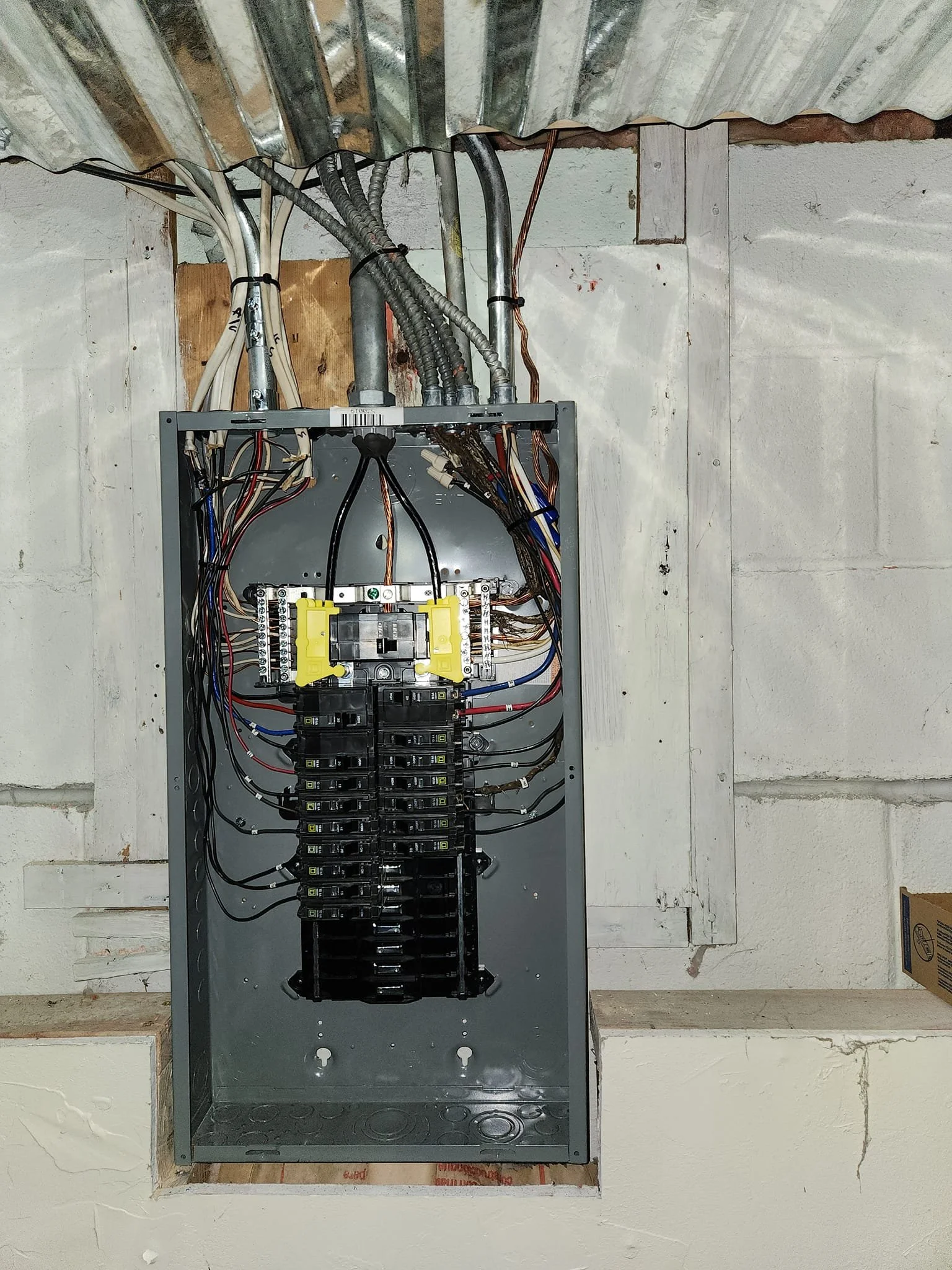 A gray electrical breaker box is open, revealing numerous black circuit breakers and colorful wires connected to terminals. The box is mounted on a concrete wall in what appears to be a basement or utility room, with a corrugated metal ceiling and wooden framing above. The wiring is neatly organized with zip ties. Some text is visible on a sticker on the top right of the box, along with a barcode.