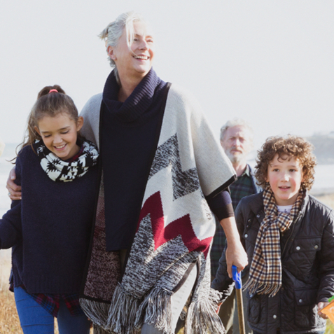 A joyful family strolls along a grassy beach path, with the ocean in the background. A young girl holds a fishing net, smiling alongside an older woman.