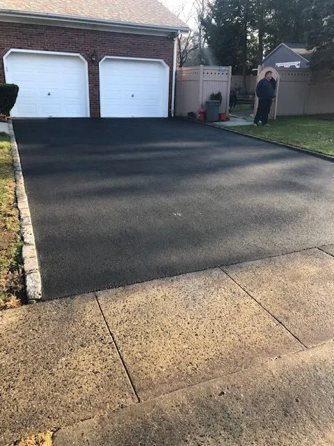 A newly paved asphalt driveway in front of a brick house with two white garage doors. A person stands in the background next to a light brown fence. Concrete sidewalk is in the foreground.