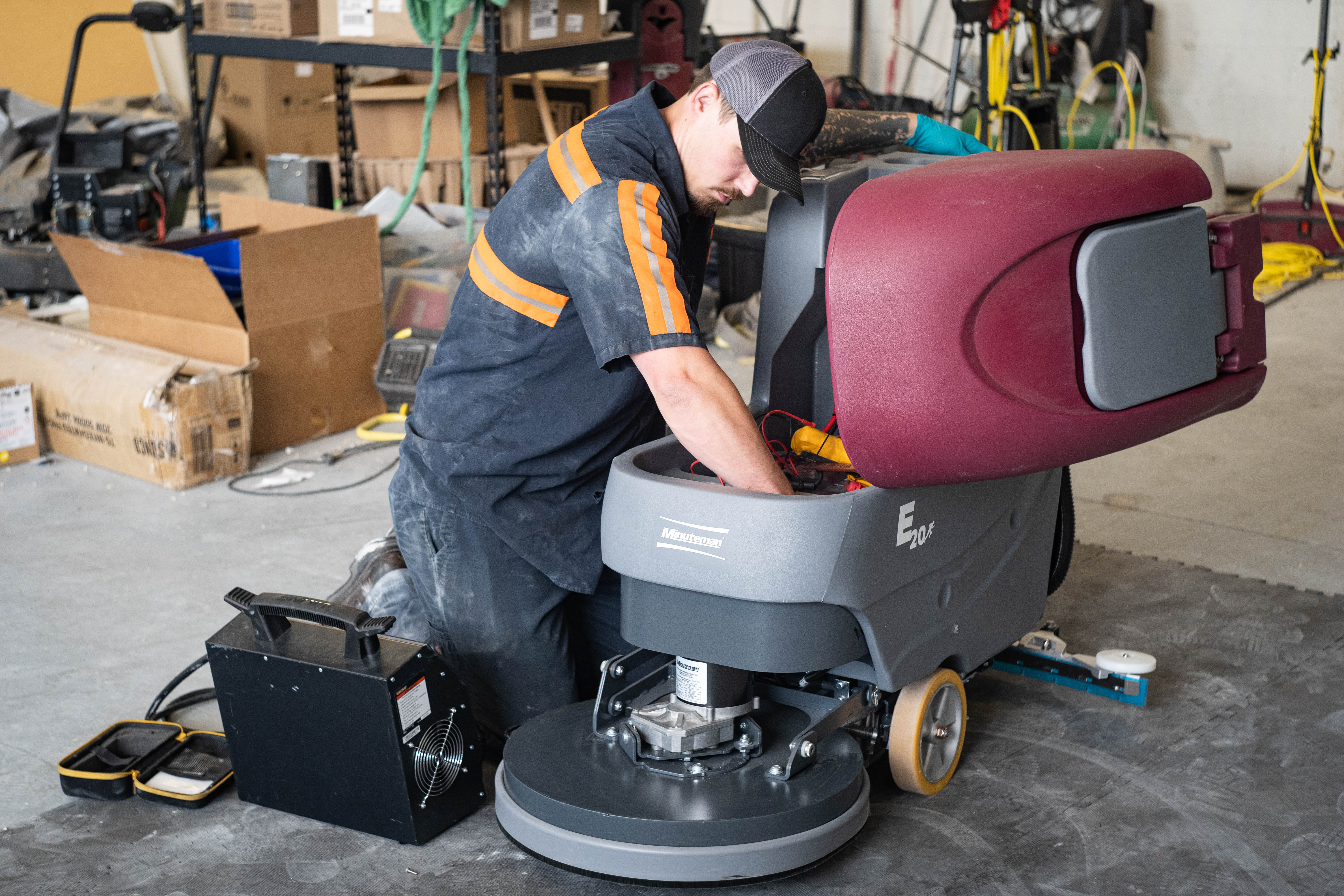 A professional WaySolutions field technician dives deep into the internal motor and battery compartment of a Minuteman E20 walk-behind floor scrubber. Equipped with professional diagnostic gear like a heavy-duty load tester, our technicians in Layton, Utah, ensure your commercial cleaning equipment maintains maximum power and scrubbing efficiency throughout its lifespan.