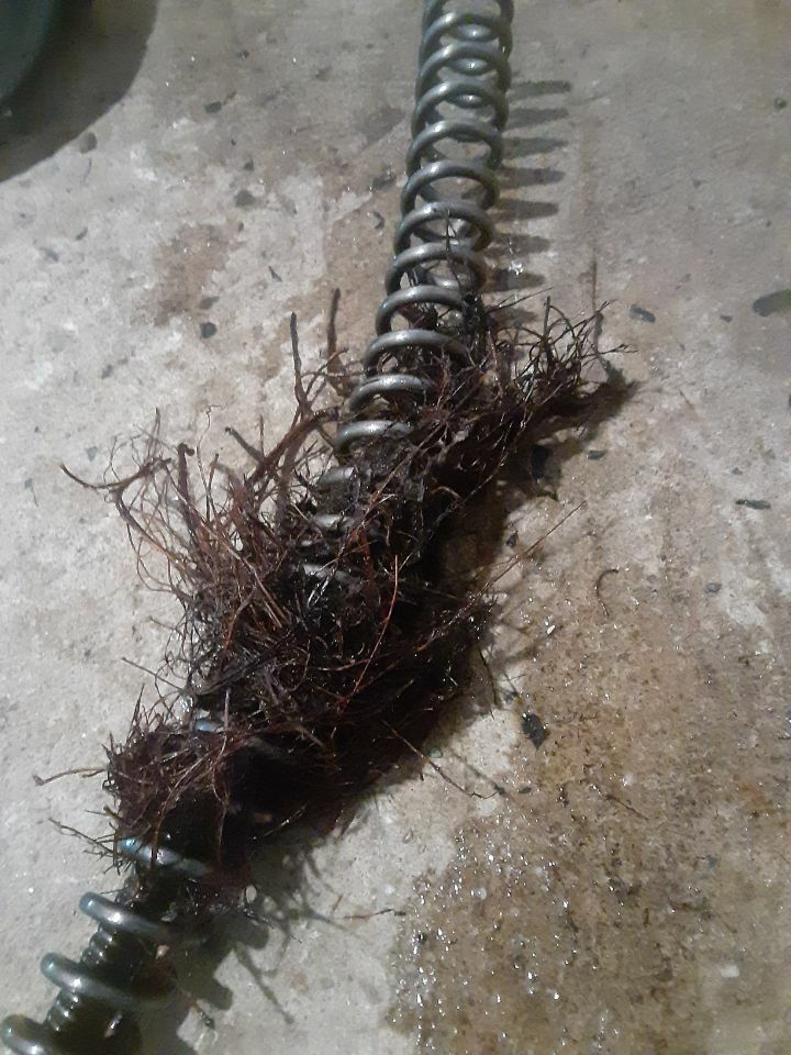 Close-up of a drain auger pulled from a pipe, wrapped with tangled hair and debris, demonstrating a severe clog. The wet surface highlights recent drain cleaning work and blockage removal.