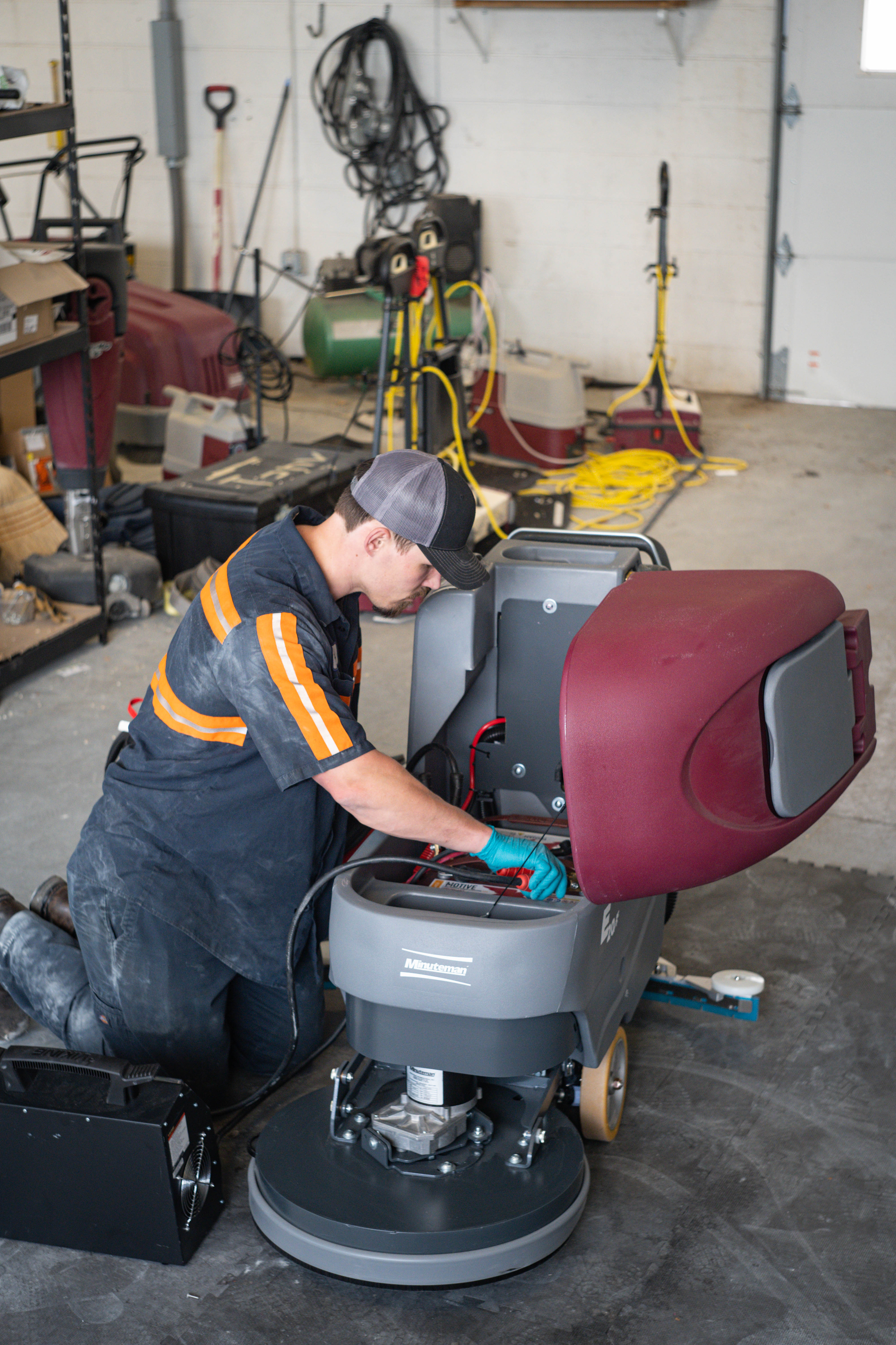 WaySolutions field technician Kaden kneels to inspect the internal battery compartment of a Minuteman E20 walk-behind floor scrubber. Serving commercial operations in Layton, Utah, our maintenance experts perform deep core power inspections to ensure your fleet's batteries and wiring remain secure and reliable.