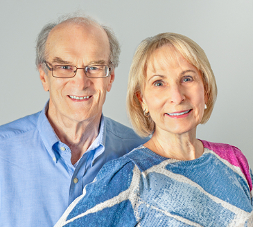 Studio headshot of a smiling senior man with glasses and a blue button-down shirt standing alongside a woman with blonde hair in a patterned sweater.