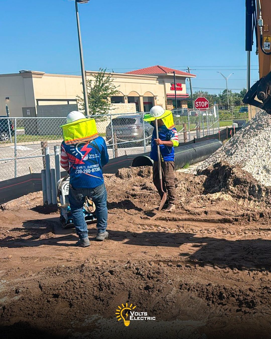 Construction workers wearing protective gear compacting soil and preparing trenches for underground electrical conduit installation at a commercial site, working near fencing, machinery, and nearby buildings.