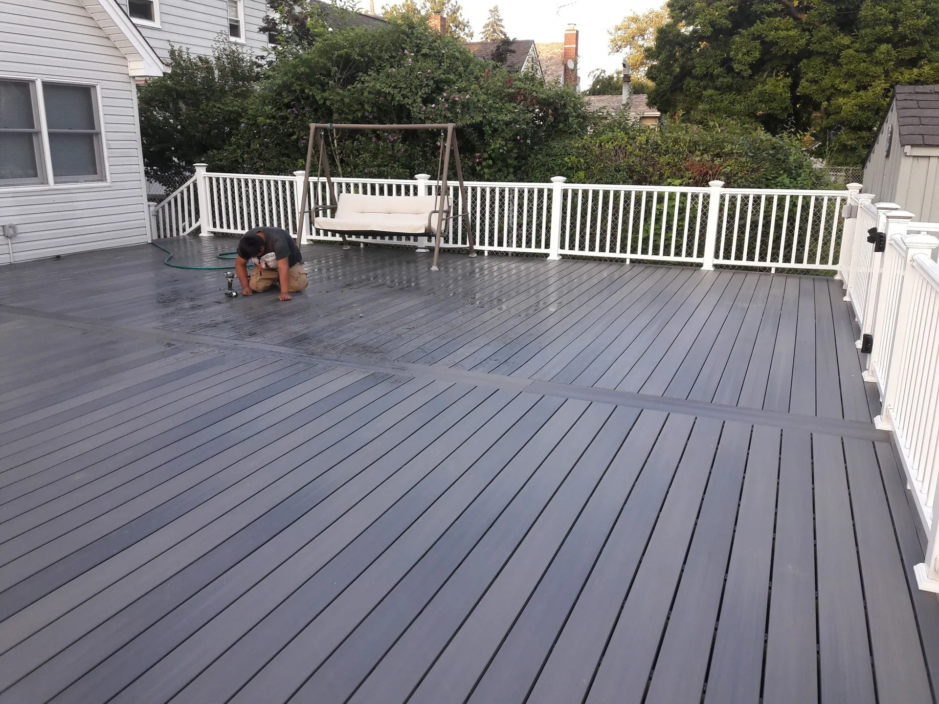 Large finished composite deck with white railing surrounds a backyard area. A contractor works on final adjustments, showcasing durable materials, spacious design, and professional outdoor construction craftsmanship.