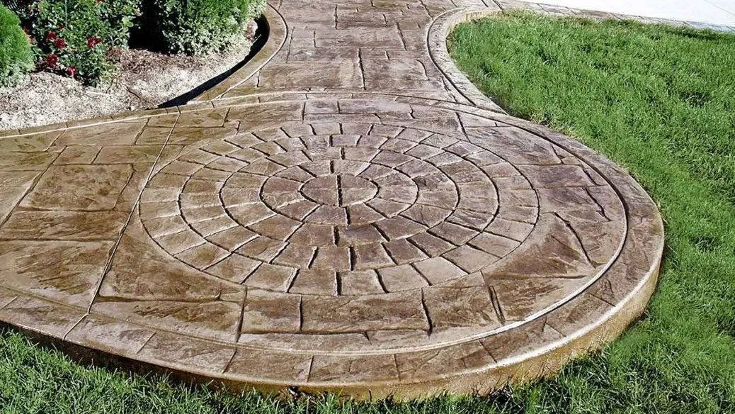 A decorative stamped concrete walkway with a circular pattern in the center, surrounded by cobblestone-style stamping, curves through lush green grass and a garden bed with red flowers.