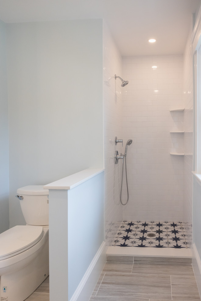 A modern bathroom featuring a white toilet to the left, separated by a light blue partition from a white tiled shower with a starburst pattern on the floor. The shower has a chrome showerhead and handheld shower wand. The floor outside the shower is tiled with a wood-grain pattern. The overall aesthetic is clean and bright, with subtle pops of blue.