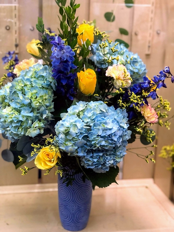 Blue Skies are the best part of a day, so why keep it outside? Sweet blue Hydrangeas accented with golden Roses and deep blue Delphinium. Blue vase may vary.