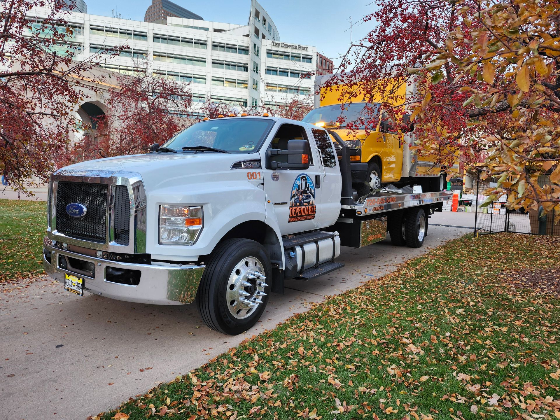 Dependable Towing & Recovery Services truck carries a yellow service vehicle in an urban setting with buildings and autumn trees, showcasing reliable city towing and professional vehicle transport services.