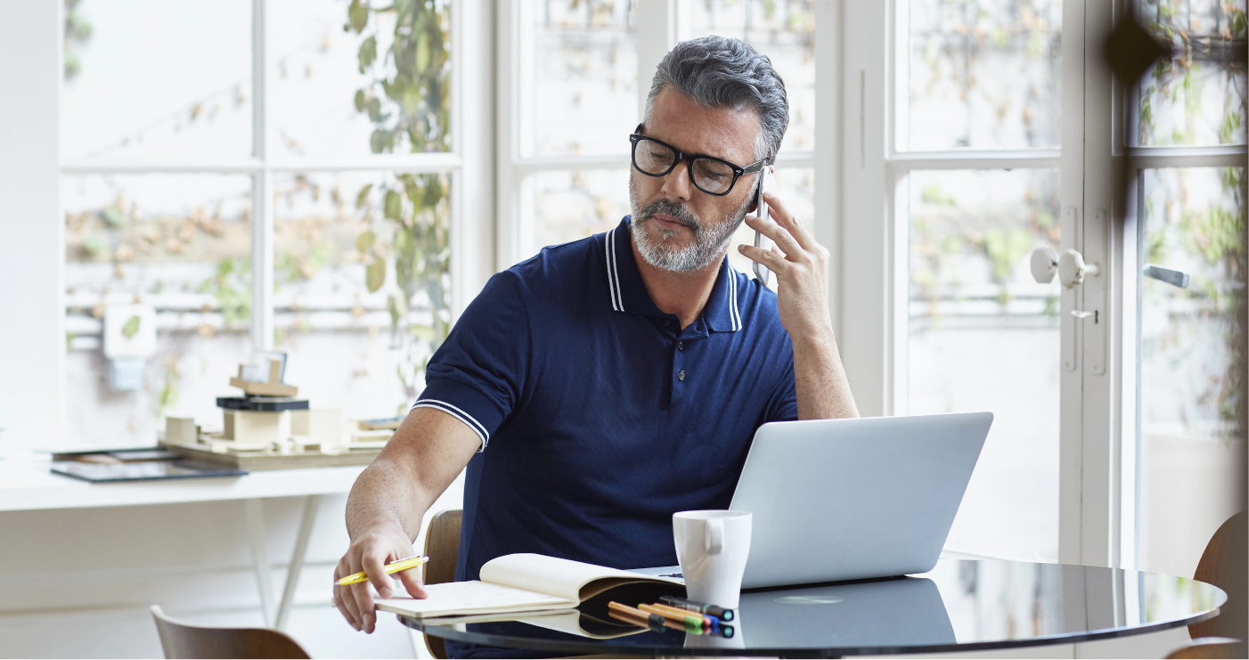 A man with graying hair and a beard is sitting at a round table and talking on his phone. He is wearing glasses and a dark blue polo shirt. On the table in front of him is an open laptop, a white mug, an open notebook, pens, and pencils. Behind him is a large window with a view of foliage. To the left of the window, there is a white desk with architectural models on it.