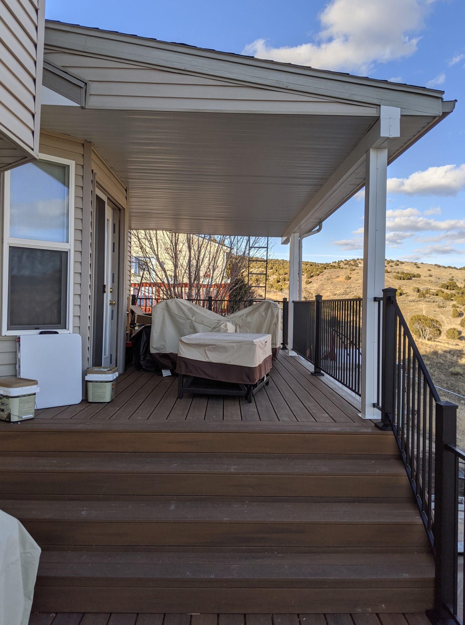 Take a step up into complete comfort with this covered composite deck by DeckPro LLC. This design seamlessly connects a lower patio to a covered upper-level living space via a wide staircase, providing an expansive area for relaxing and entertaining. The integrated roof cover offers reliable shelter from the elements, while sleek black metal railings define the perimeter, blending modern safety with a rustic mountain-side view.