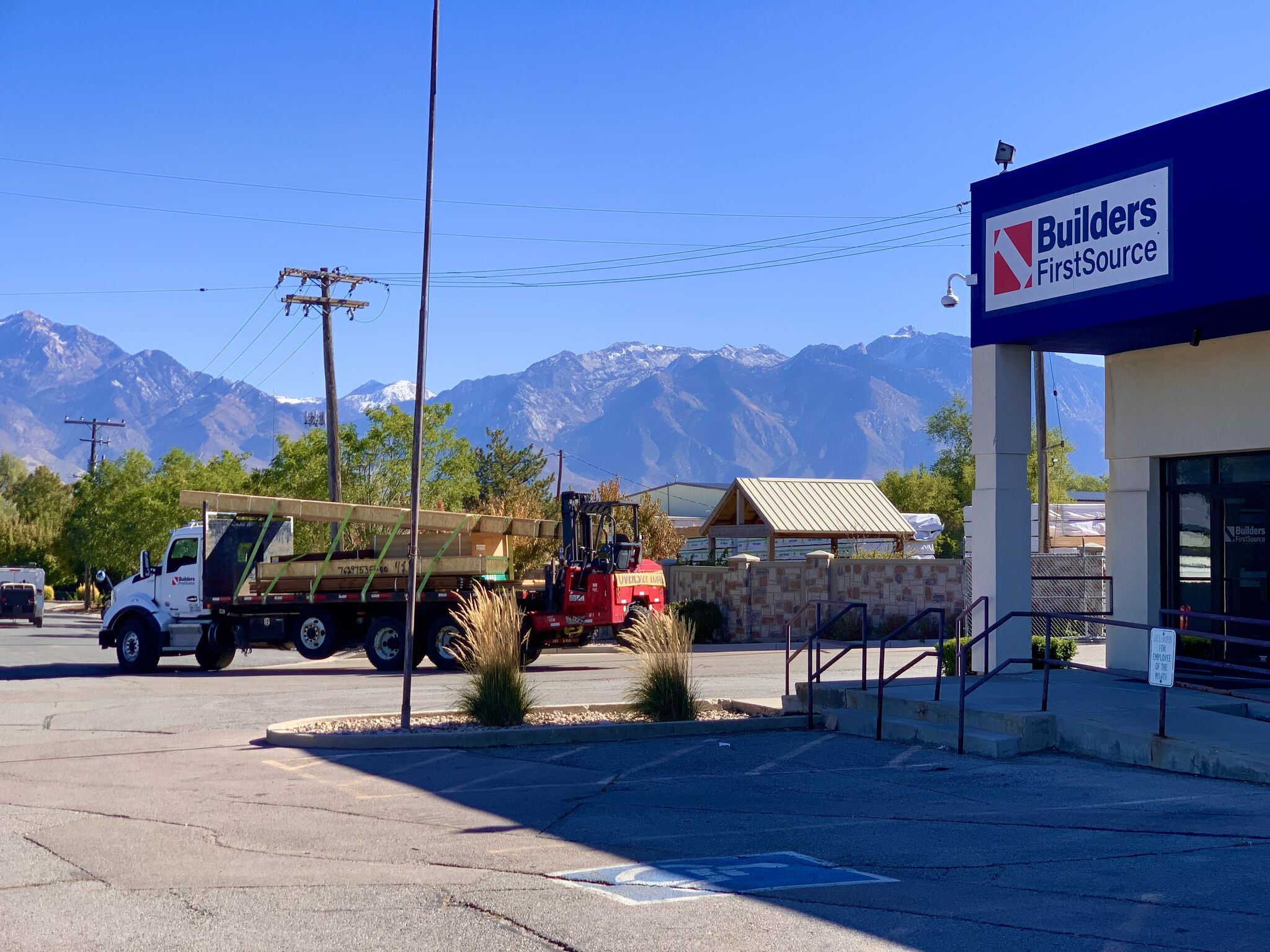 Builders FirstSource location with a delivery truck and forklift in front of a stunning mountain backdrop under a clear blue sky.