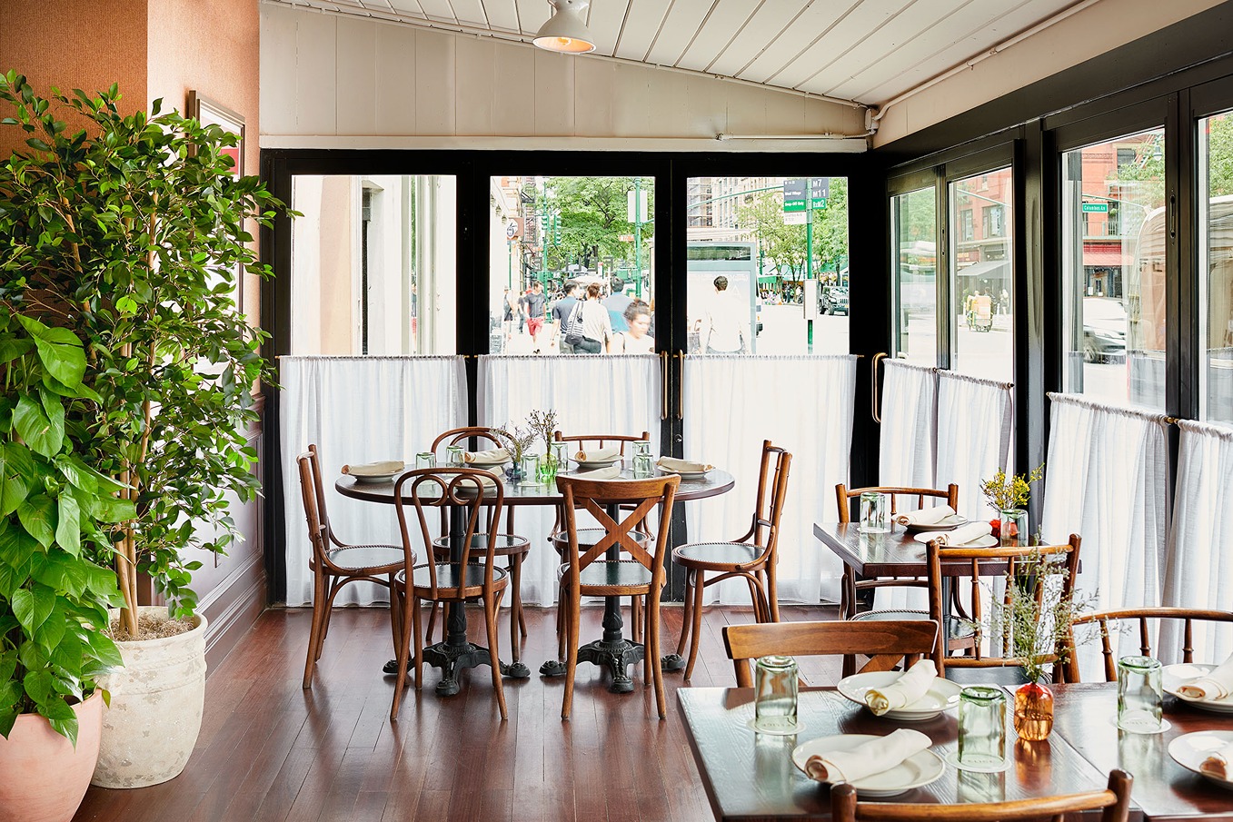 Sunlit restaurant interior with wooden tables and chairs. Large black-framed windows with white caf&eacute; curtains offer a view of a busy sidewalk and green trees outside.