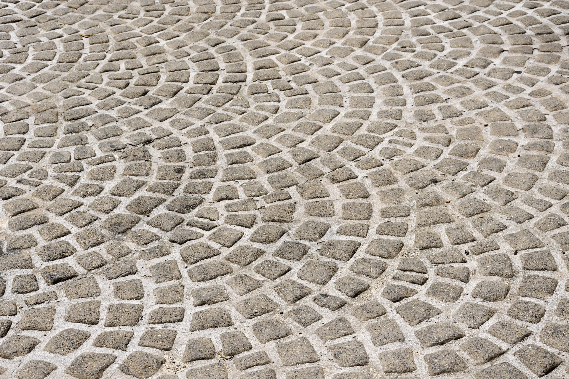 A detailed, close-up shot of a cobblestone pathway or plaza, laid in a fan or semi-circular pattern. The stones are irregular in shape and size, with visible texture and grain, and are set in sand or gravel. Sunlight casts subtle shadows, highlighting the contours of the paving.