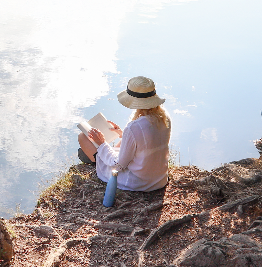 A woman reading a book by a calm body of water.