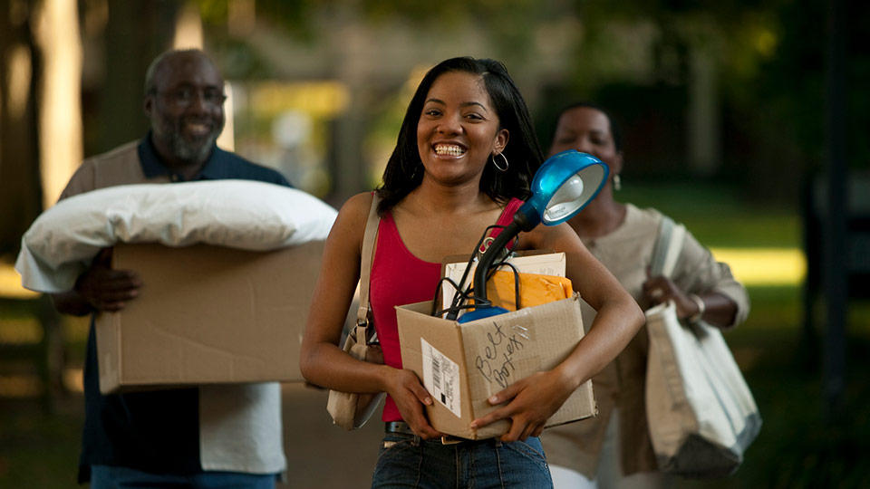 A smiling student carries a box from her dorm with her parents carrying boxes behind her.