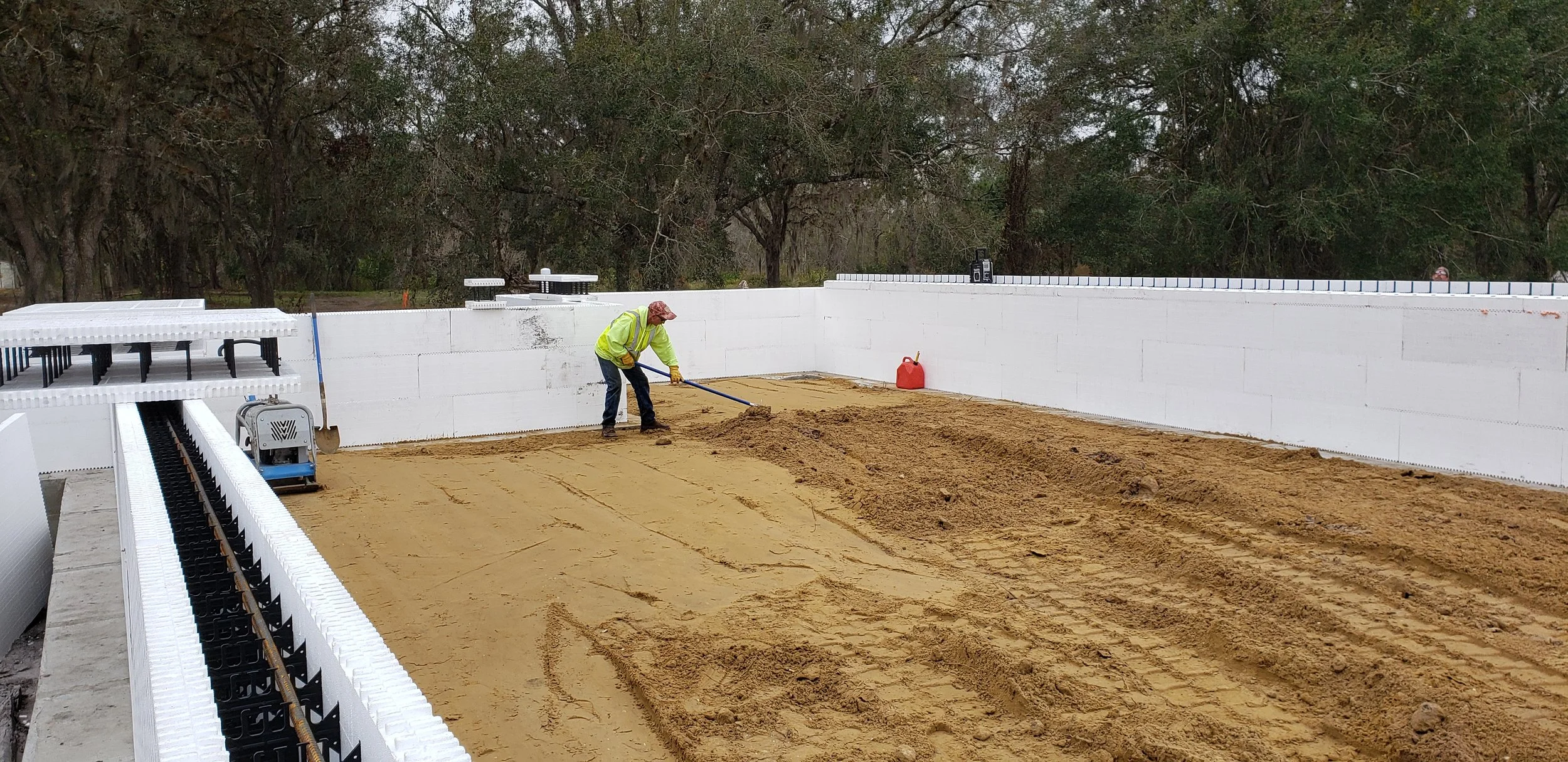A construction worker in a high-visibility vest and hard hat uses a shovel to move sand inside a fou 