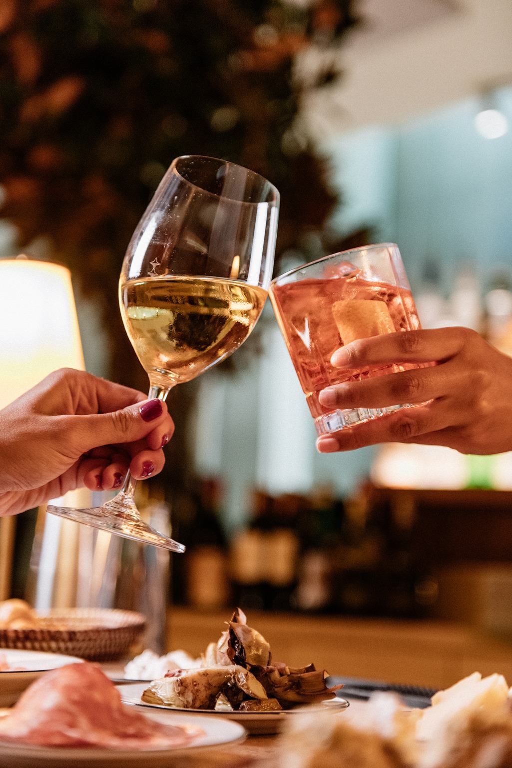 A warm, candid shot of two hands raising a glass of white wine and an orange cocktail in a "cheers" over a table of Italian appetizers and cured meats.