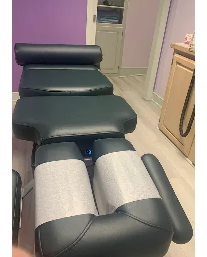 It&rsquo;s an interior photo of a treatment room featuring a black padded chiropractic/therapy table with segmented cushions and a raised headrest. Two light gray fabric pads are positioned over the leg/foot section, and there are padded side supports/armrests.
The room has lavender/purple walls, light-colored flooring, and a doorway/opening leading toward a small back area. On the right side, there&rsquo;s a wood cabinet/counter with a coiled cord/handheld device hanging down, giving the space a clean, clinical feel.