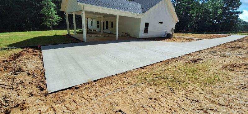 Newly poured concrete driveway extending from covered carport beside white house, surrounded by graded soil and lawn, with smooth finish and clean edges in residential setting.