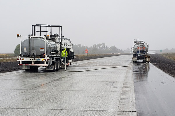 A wide-angle perspective photograph looking down a newly ground concrete highway on a overcast, foggy, or rainy day. The wet concrete surface reflects the dull sky. In the foreground-left, a massive truck-mounted DSI diamond grinding rig (white with silver tankers) is operational, connected via a hose to a second DSI slurry tanker truck parked on the shoulder-right. The shot captures the vast scale of heavy highway grinding on a public works project.
