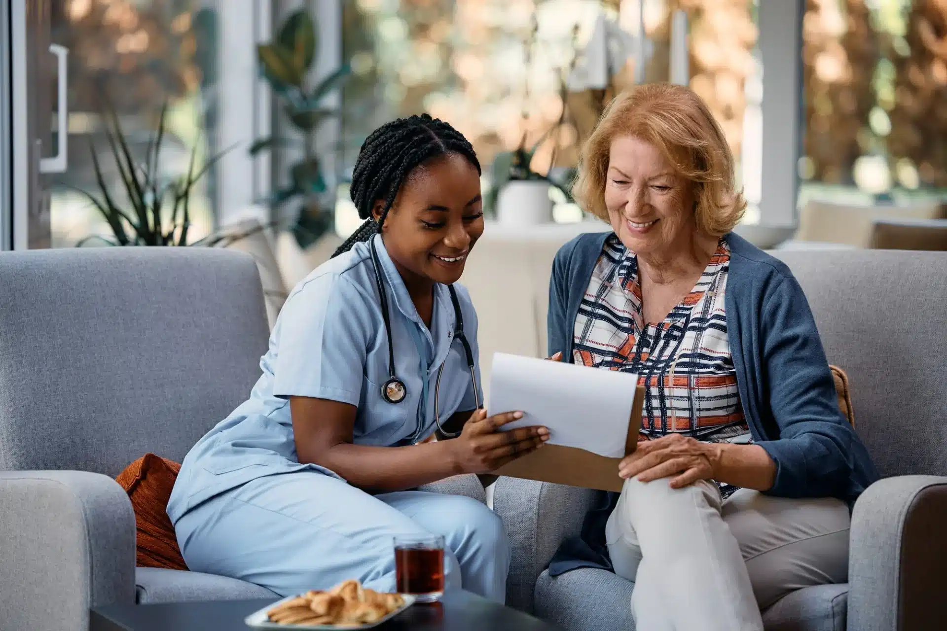 A nurse in light blue scrubs and a stethoscope around her neck sits on a sofa and smiles while looking at a document with an elderly woman. The woman is wearing a plaid shirt and a blue cardigan and also smiles while looking at the document. A cup of tea and a plate of cookies are on the coffee table in front of them. The background is softly blurred, suggesting an indoor setting like a waiting room or a comfortable living area.