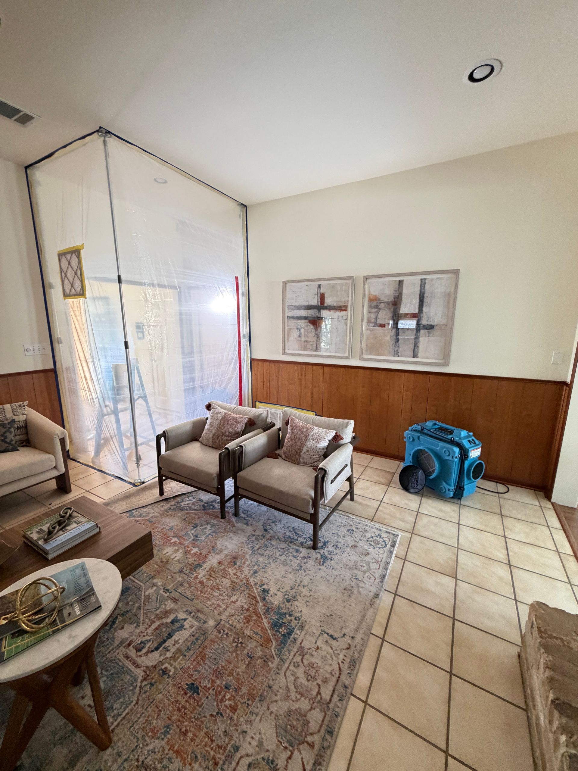 A furnished living room is partially sealed with clear plastic containment barriers while a blue air 
