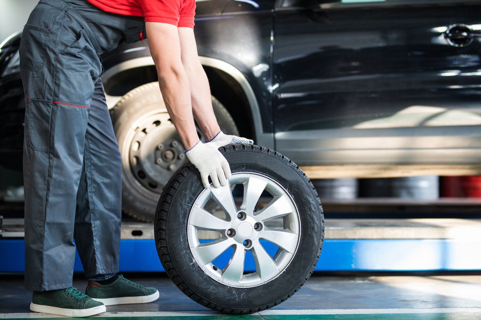A mechanic in grey overalls and a red shirt rolls a tire across a garage floor toward a black car on 