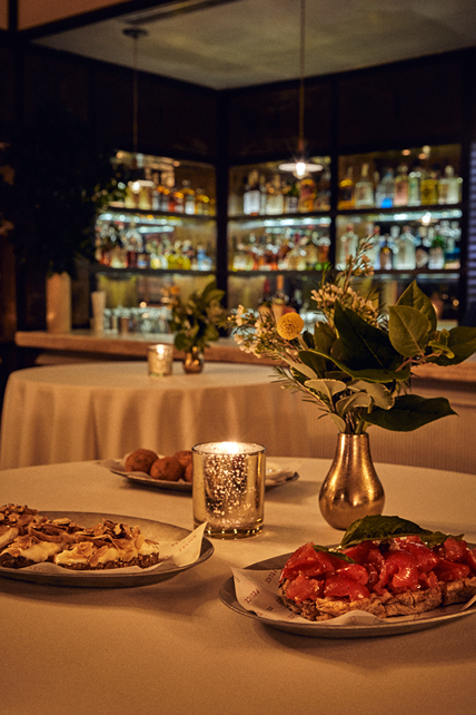 Plates of bruschetta and small bites sit on a white tablecloth lit by a shimmering candle and a small floral arrangement. In the blurred background, a well-lit bar displays a variety of spirits.