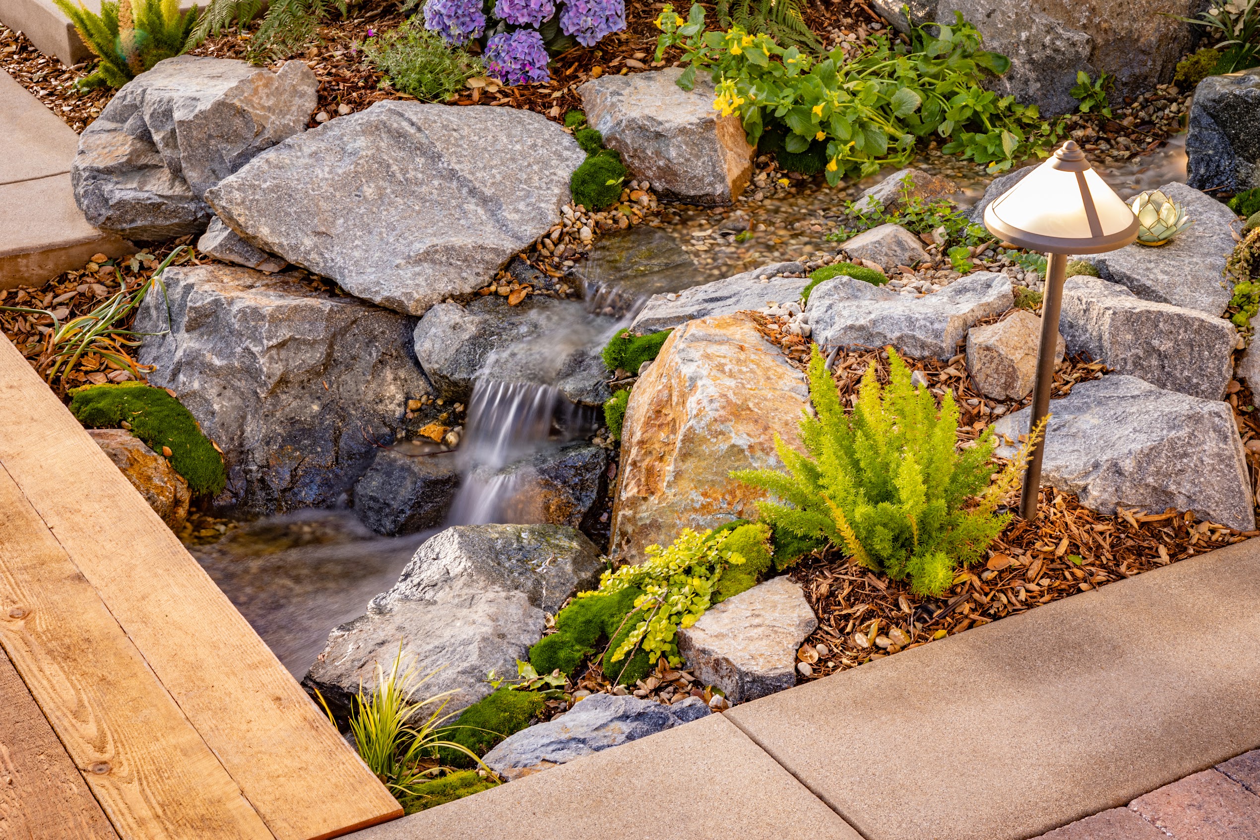 Serene landscaped garden stream cascades over rocks beside lush ferns, purple flowers, a wooden bridge, and a glowing path lamp.