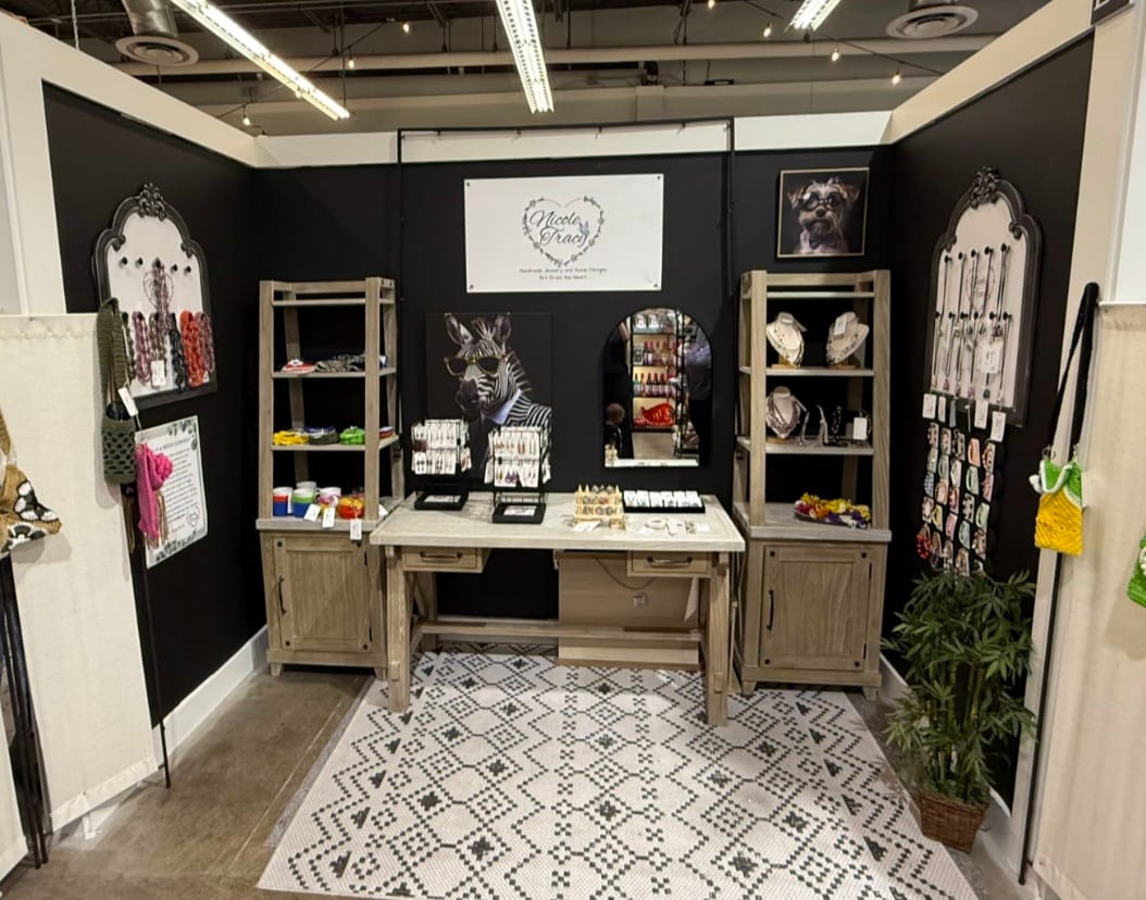 A display booth at a boutique featuring handmade jewelry and home designs. The booth has black walls and a light-colored rug with a geometric pattern. It showcases various jewelry pieces on shelving units and display stands, along with framed artwork, including a dog wearing glasses and a stylized zebra. A wooden desk is placed in the center, offering a surface for displaying items. There are also decorative items and plants.