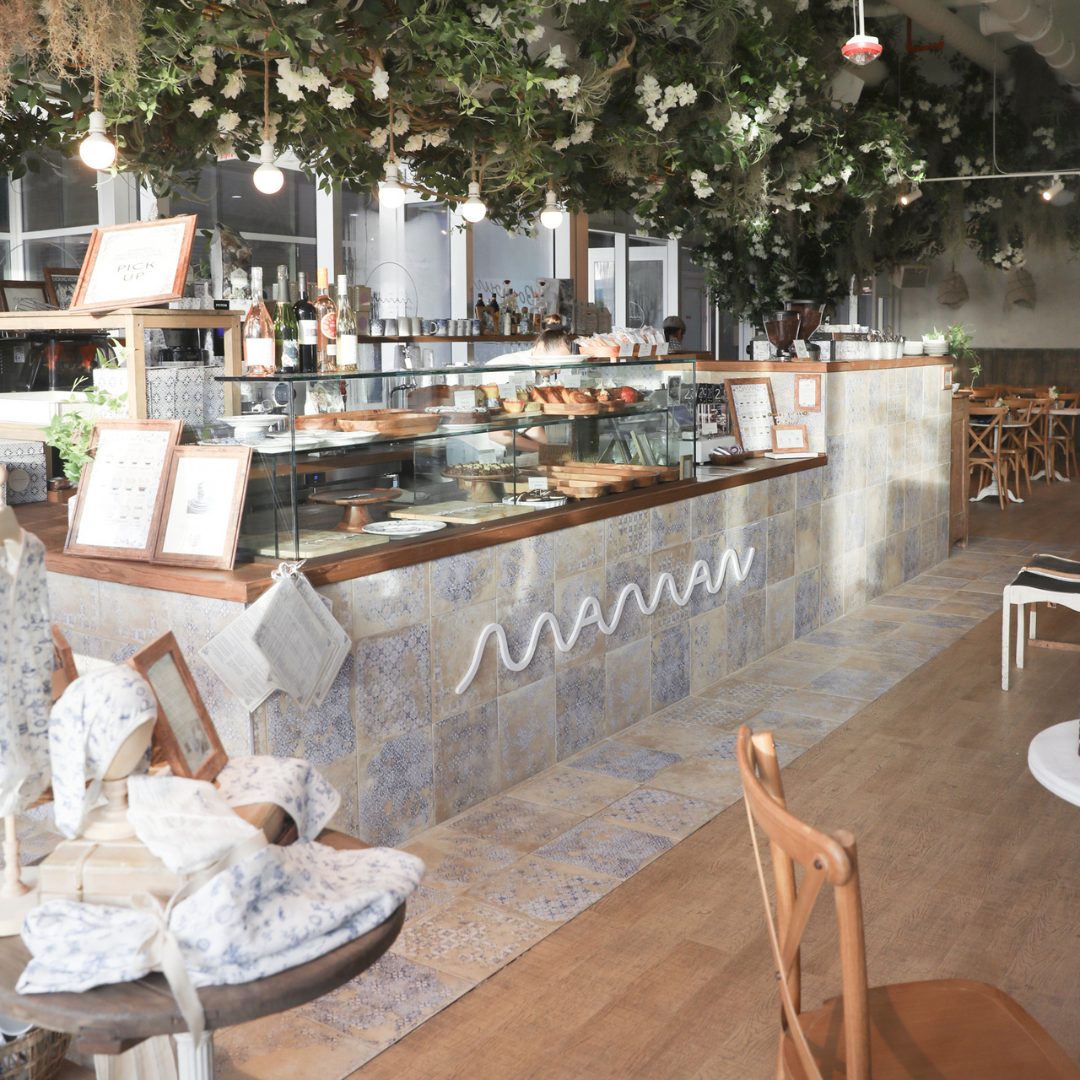 The café unfolds from the pastry counter into the dining room, where wood, stone, greenery, and natural light blend with shelves of pastries and everyday movement.