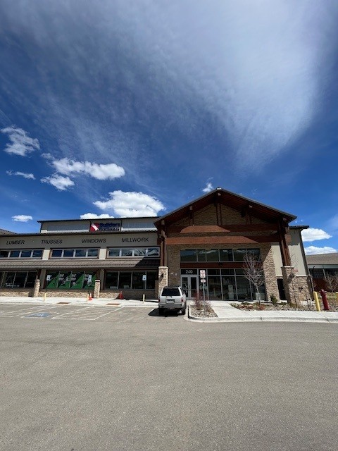 Exterior view of a Builders FirstSource building materials showroom with a modern peaked entrance, large glass storefront, and signage for lumber, trusses, windows, and millwork under a clear blue sky.