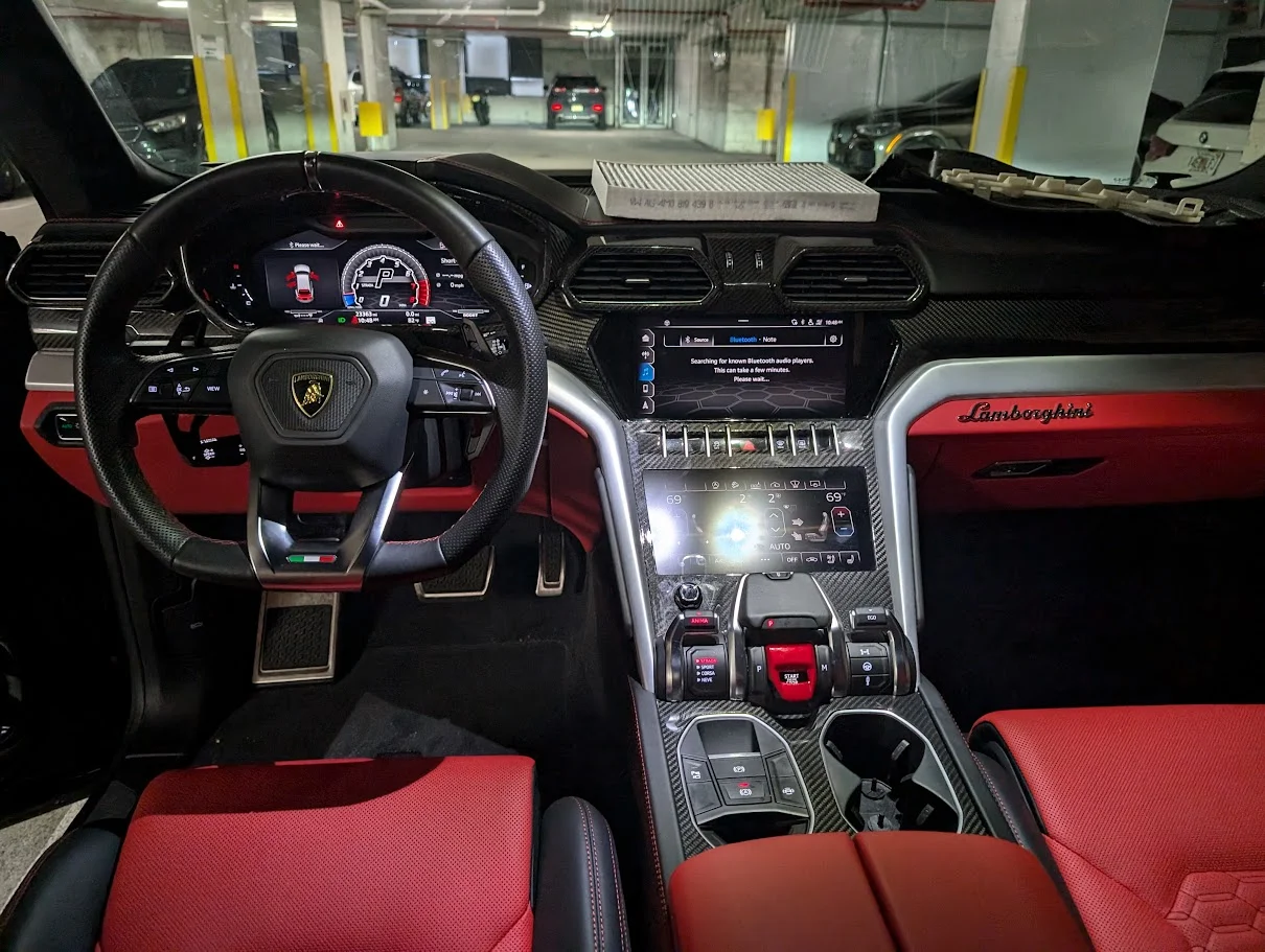 The interior of a luxury car, likely a Lamborghini Urus, is shown from the driver's perspective. The dashboard features a large digital instrument cluster, a central infotainment screen, and various controls. The seats and dashboard accents are red, contrasted with black leather and carbon fiber trim. The car is parked in what appears to be an indoor parking garage.