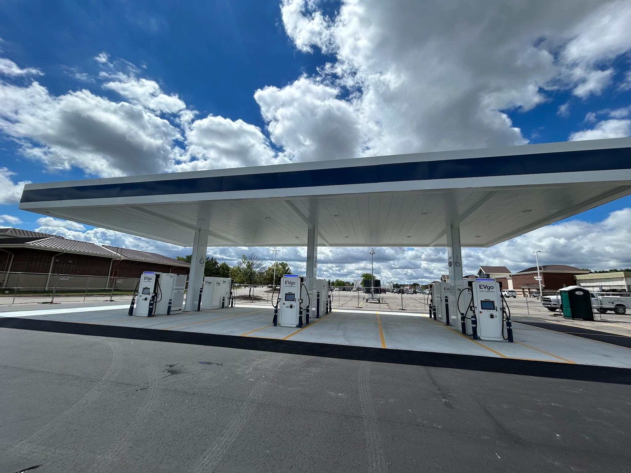 EVgo fast charging station with multiple fast chargers arranged in rows beneath a large white canopy with navy blue trim, set against a bright blue sky with scattered clouds.