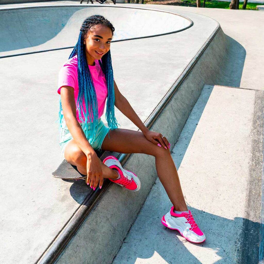 A young Black woman with bright blue box braids is sitting on a skateboard in a skate park. She is wearing a pink t-shirt, turquoise shorts, and white and pink sneakers. She is smiling at the camera. The sun is shining, casting shadows on the concrete.