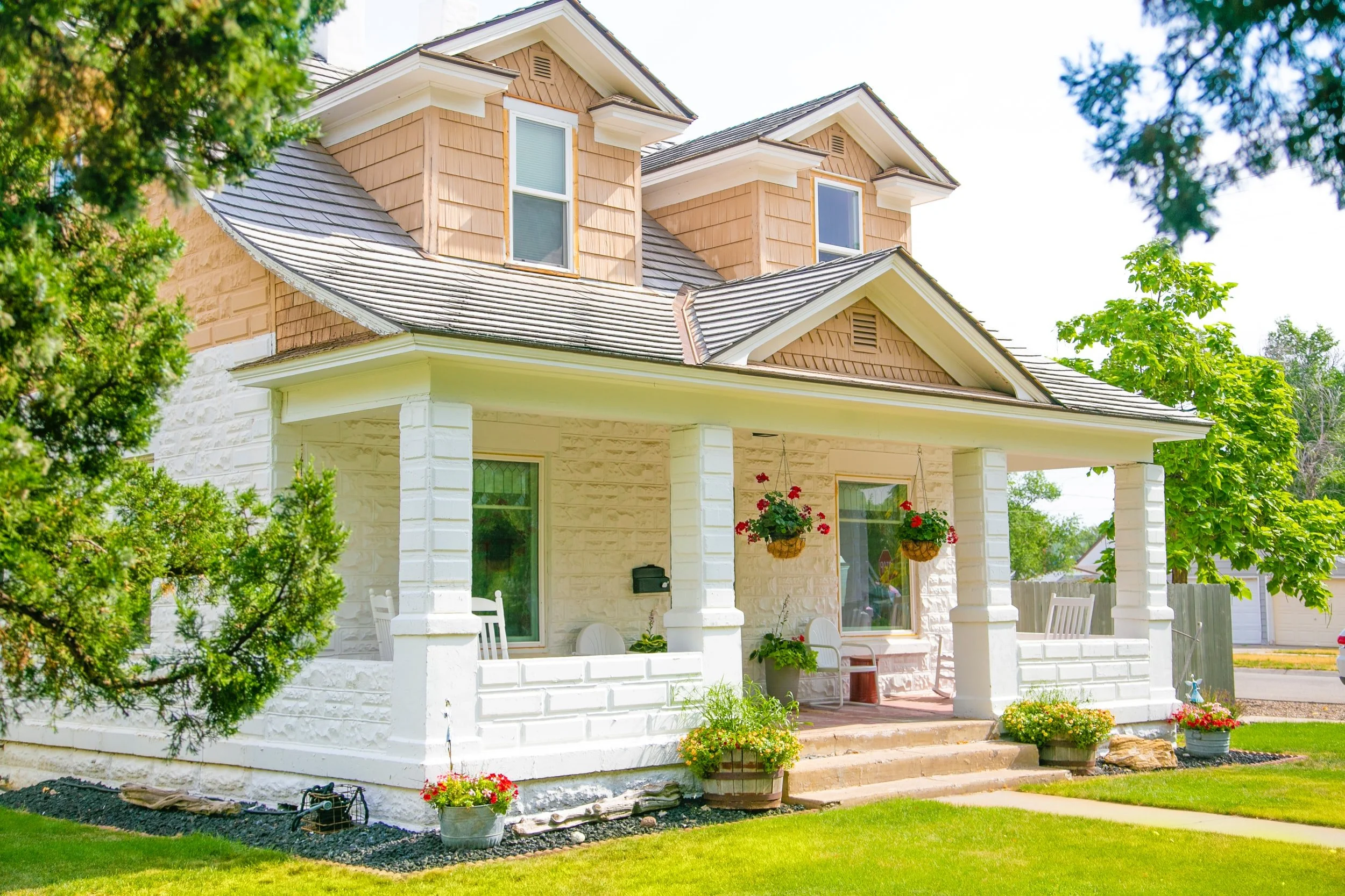 A charming two-story house with white brick and tan shingle siding sits on a green lawn under a bright sky. The house features a wraparound porch with white columns, hanging flower baskets with red geraniums, and outdoor chairs. Landscaping includes potted plants and decorative rocks. In the background, trees and a wooden fence are visible, along with garage doors.