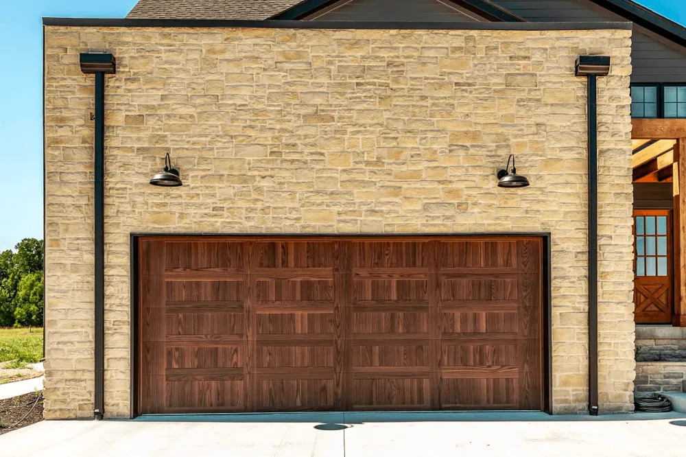 A stone garage building features a rich wood double garage door with horizontal detailing, framed by dark trim, exterior wall lights, and warm natural stone masonry creating timeless curb appeal.