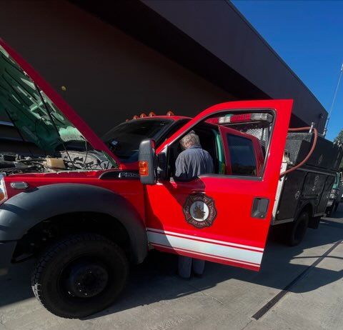 Red service truck with its hood and driver door open while a technician works inside the cab, showing heavy-duty tires, utility body compartments, emergency-style markings, and outdoor service area nearby.