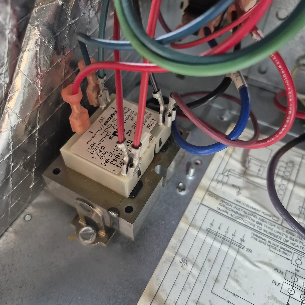A close-up shot shows a cream-colored electronic relay with red, blue, green, and black wires connected to it. The relay is mounted on a metallic bracket. The background features a textured metallic surface, likely insulation, and part of a wiring diagram on a paper sheet. The scene appears to be inside a piece of electrical equipment.