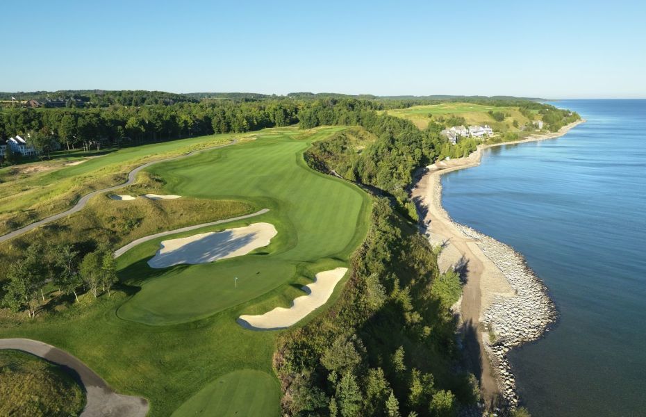An aerial drone shot shows a meticulously manicured golf course with rolling green fairways, strategically placed sand traps, and a clear blue sky overhead. The course is bordered by a lush green forest and a rocky shoreline that meets the expansive, tranquil blue water of a large lake or ocean. Several elegant houses are nestled among the trees and on the hills overlooking the water, suggesting a luxurious resort or residential community.
