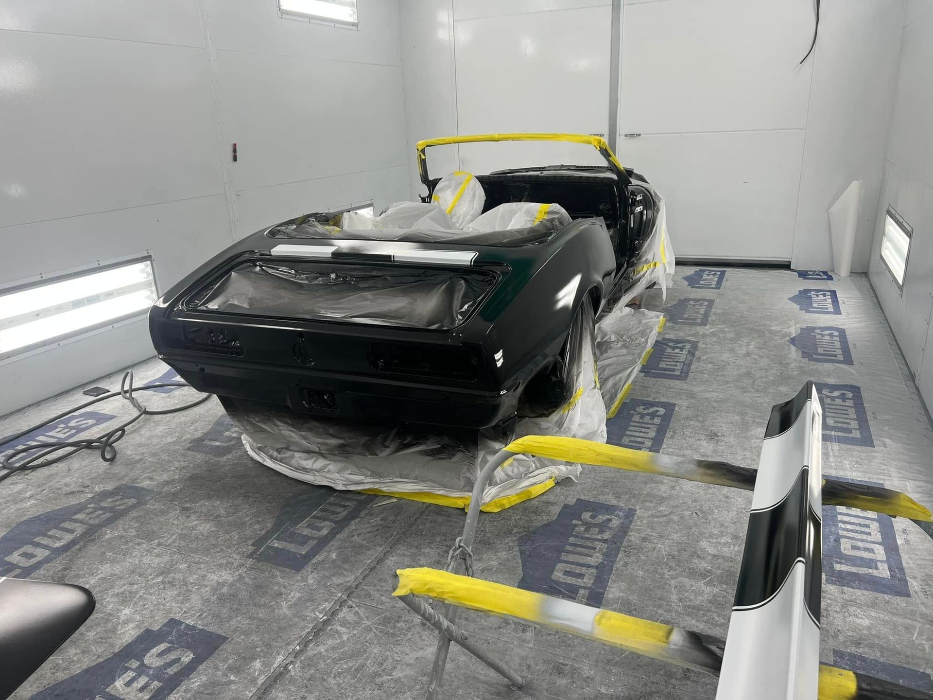 A vintage Camaro is positioned in a spray booth after paint application. Protective masking remains in place while the fresh black finish cures during restoration work.