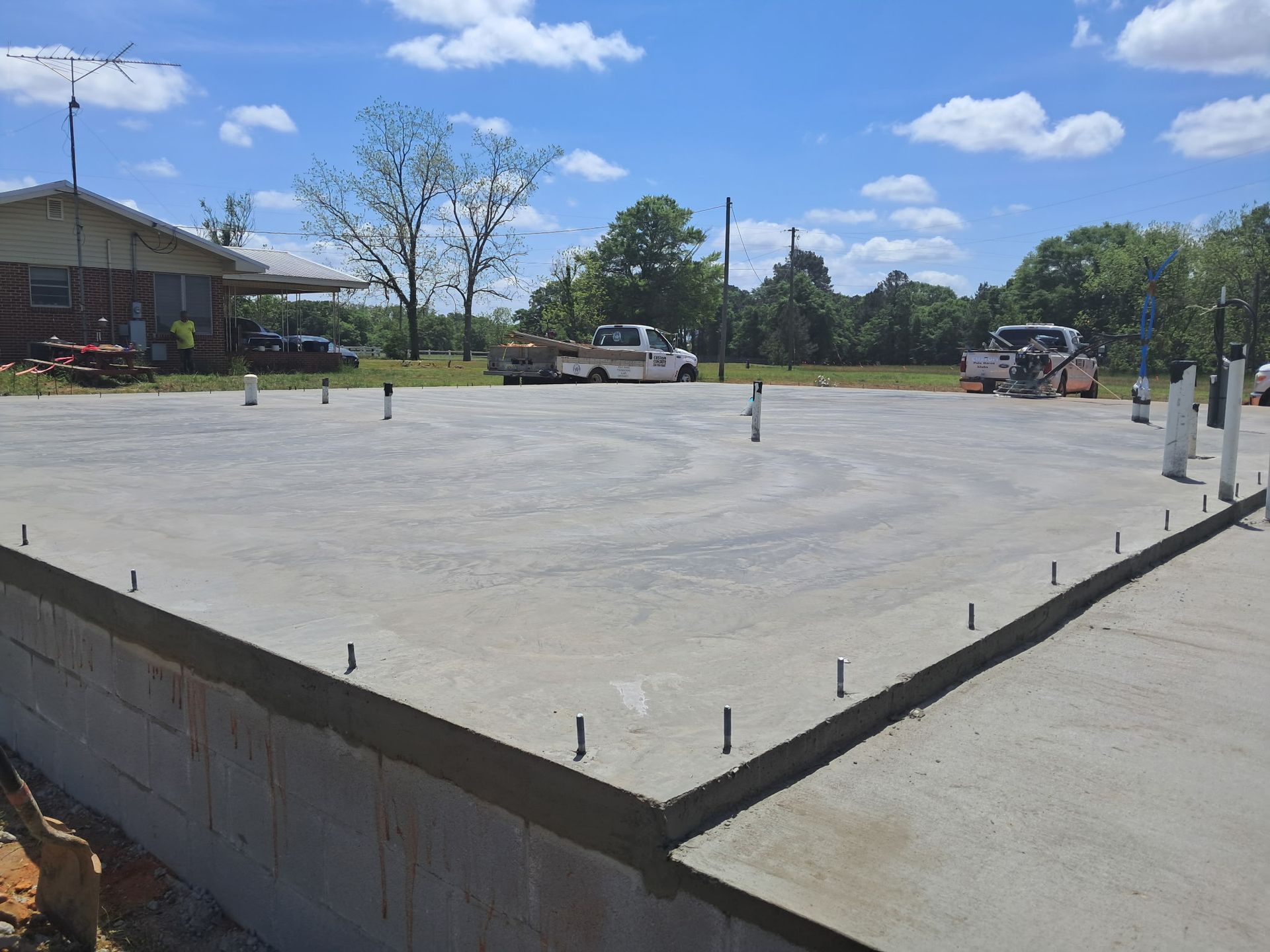 Large elevated concrete slab foundation with anchor bolts installed, work trucks and equipment nearby, rural setting with house, trees, and open sky during active construction phase.