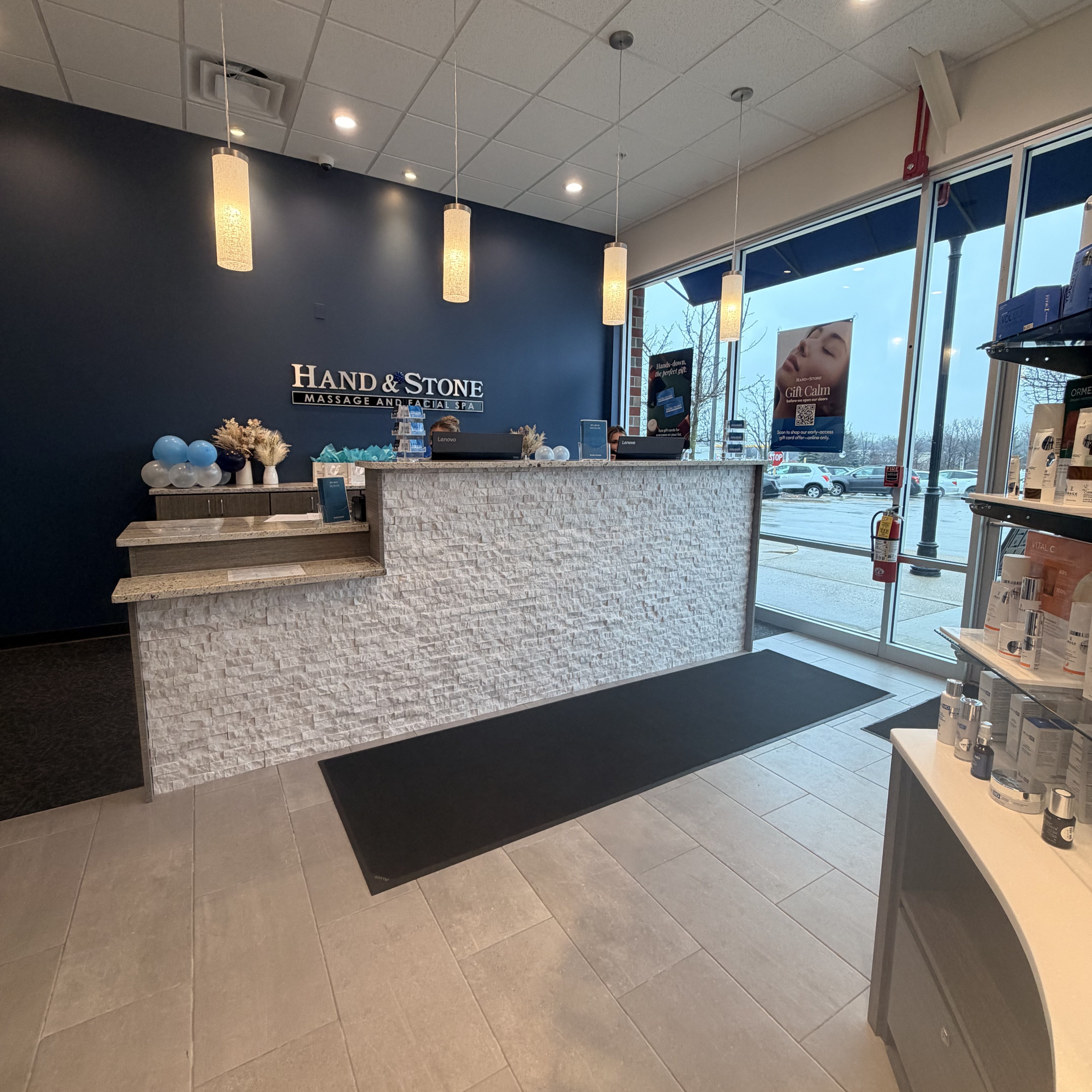 Wide-angle view of reception with Hand & Stone logo on navy blue accent wall. Stacked stone feature wall behind curved reception desk. Cylindrical pendant lights hang from white ceiling. Gray tile flooring and glass entrance doors with promotional window clings.