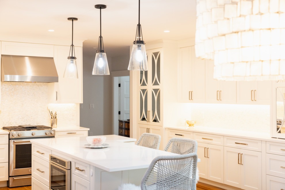 A beautifully designed modern kitchen with white cabinets, a large island with a white countertop, and stainless steel appliances. The kitchen features several pendant lights and a large, elaborate chandelier. The overall aesthetic is clean, bright, and spacious, with warm lighting enhancing the luxurious feel. There are bar stools around the island, and a glimpse into a hallway with a dark wooden dresser.