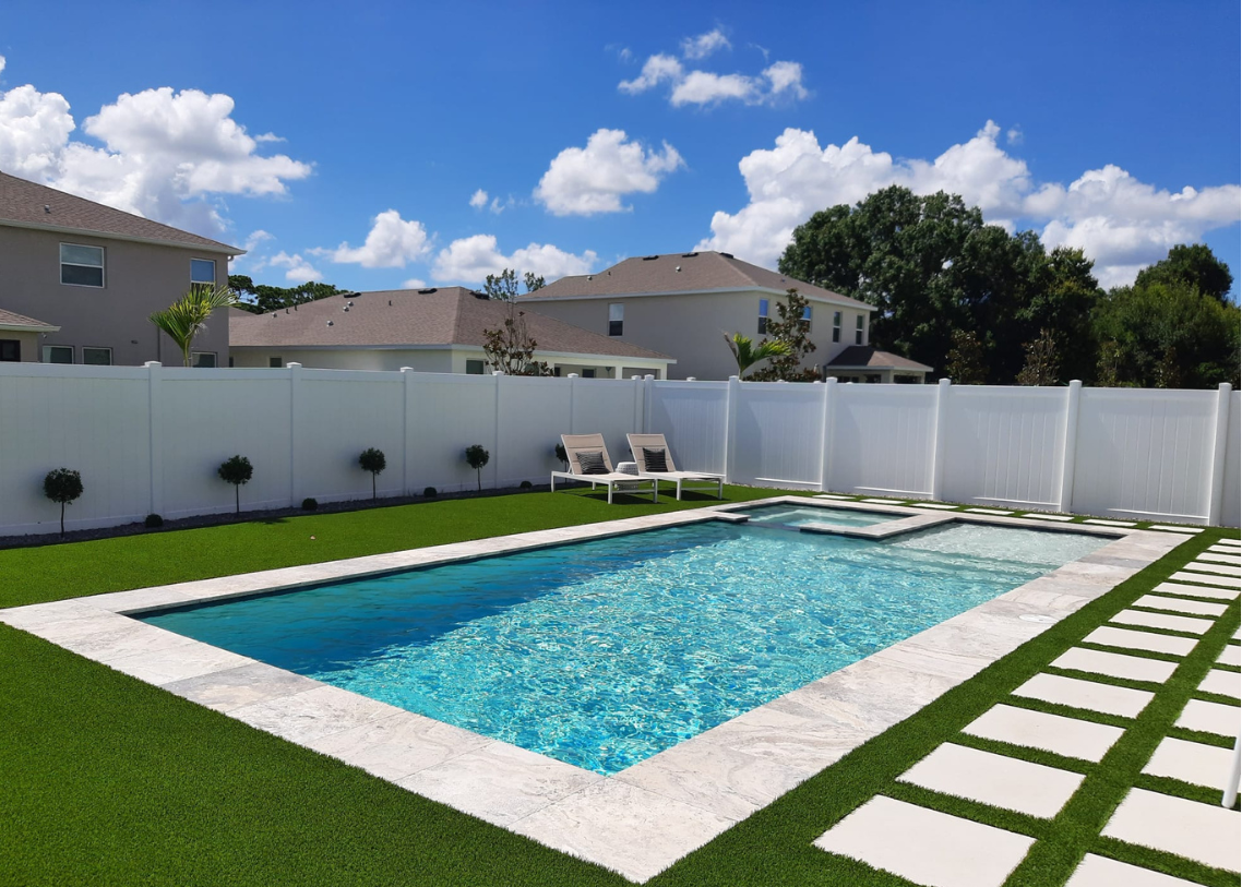 Modern backyard swimming pool with clean geometric lines, white stone decking, green turf, and a privacy fence.