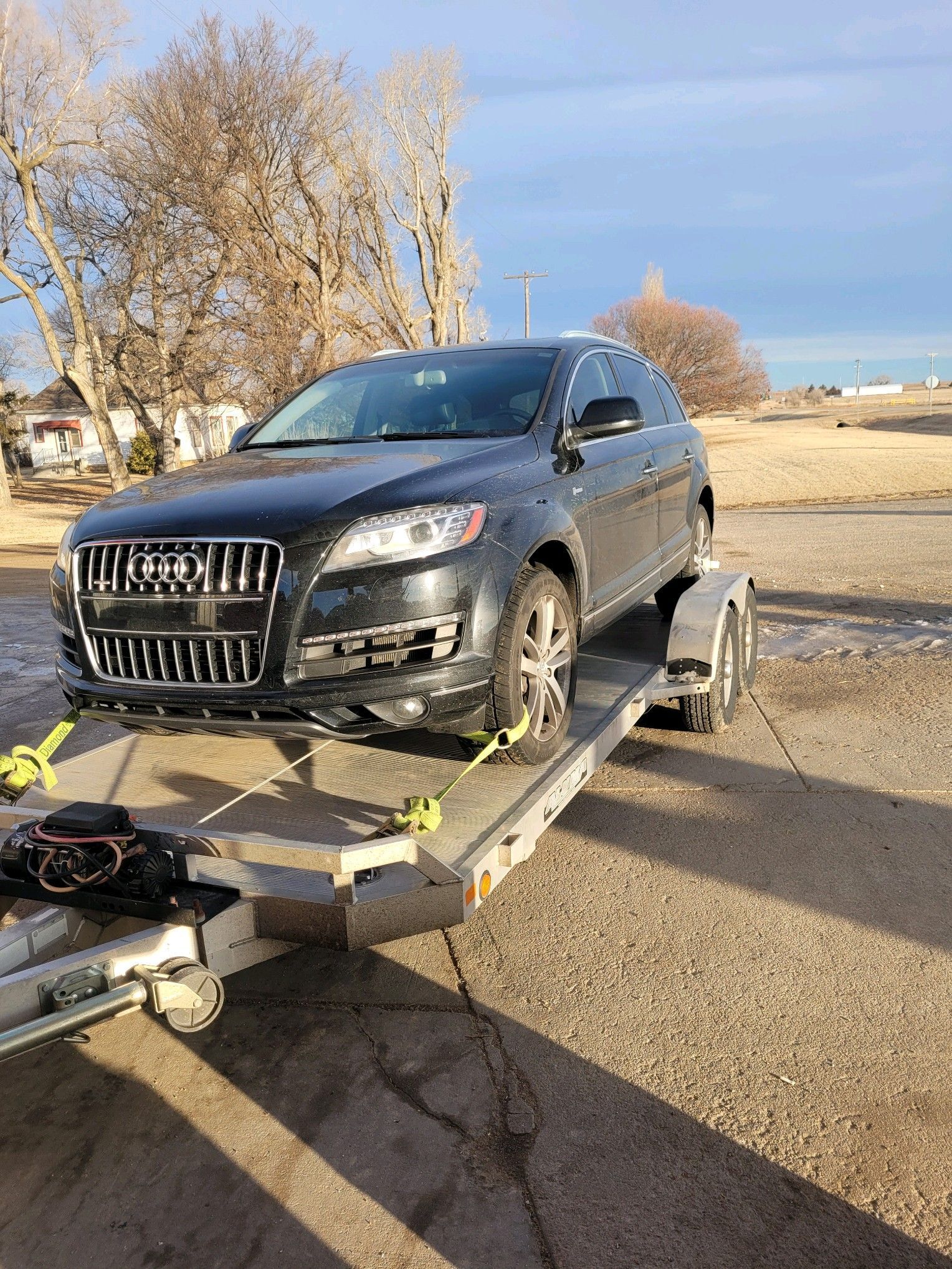 A dark gray Audi SUV rests securely on a dual axle aluminum transport trailer anchored by yellow straps, parked on a concrete driveway under a clear blue afternoon winter sky.
